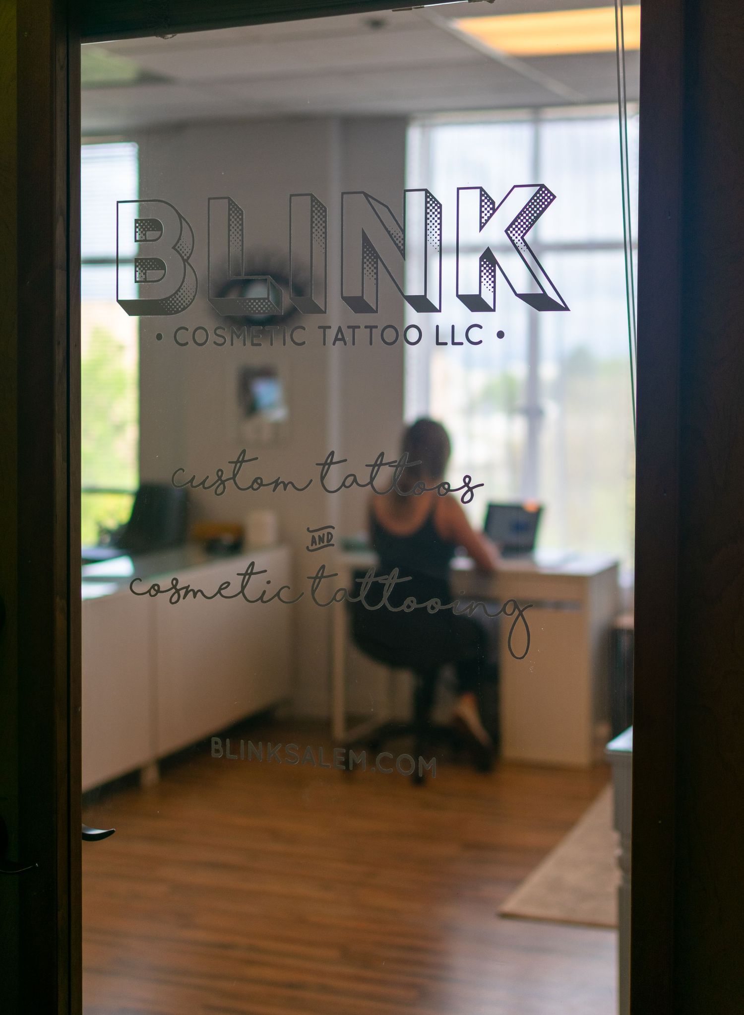 Blink Tattoo & Esthetics front door with logo showing on glass door. Amanda is sitting inside at her computer.
