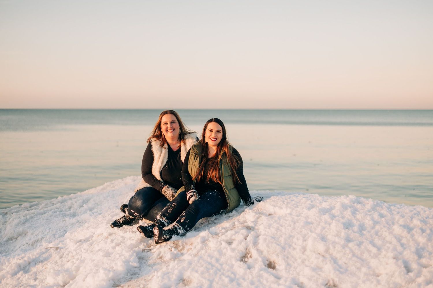 photo of two women sitting on a frozen wave on a snowy beach at sunset