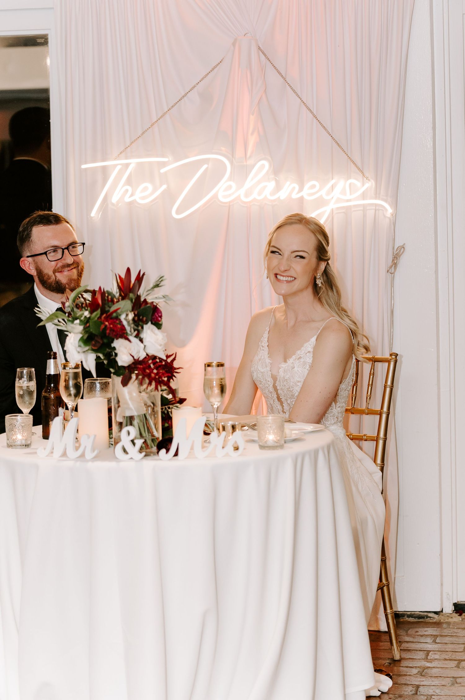 Wedding reception table decorated with white linens and floral arrangements under custom lighting.