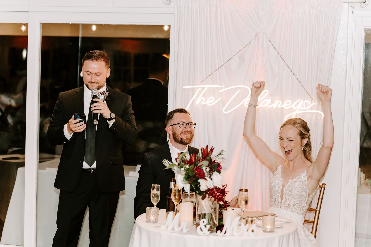 Wedding reception scene with toasts and a neon sign at the head table during an indoor celebration.