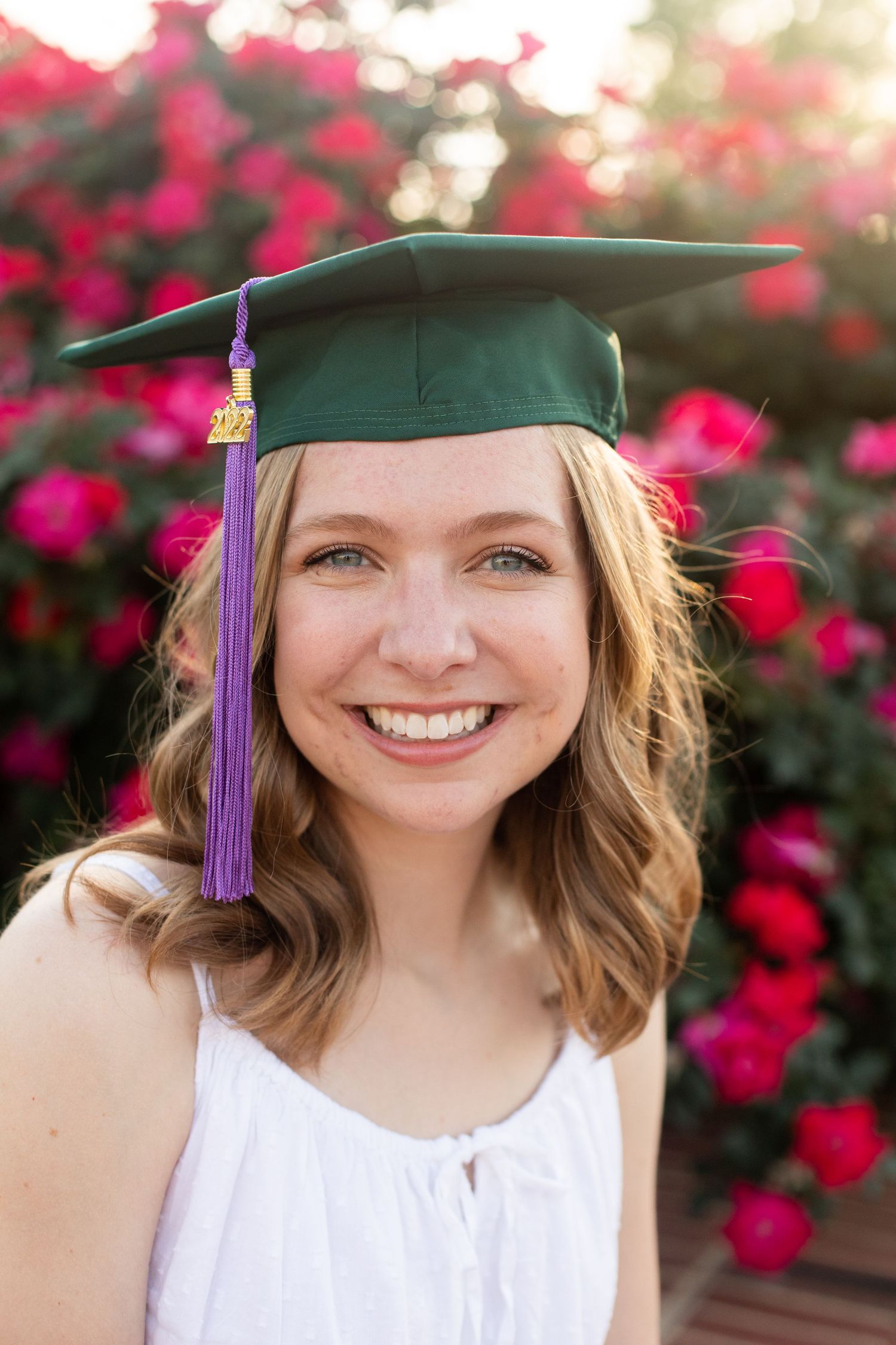 Graduate in green cap with purple tassel poses against blooming pink flowers.