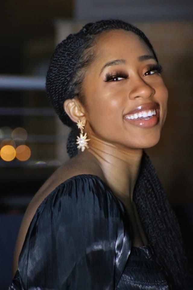 Profile portrait showcasing braided hairstyle and black evening wear against blurred lights.