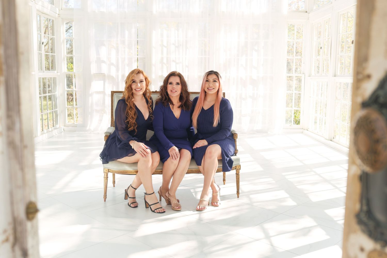Three women in navy blue dresses seated on elegant chair in bright sunlit room.