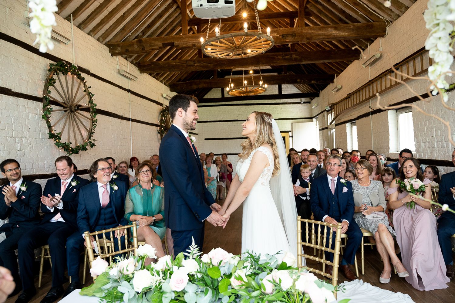 Indoor wedding ceremony with floral decorations and wooden beamed ceiling in rustic venue.