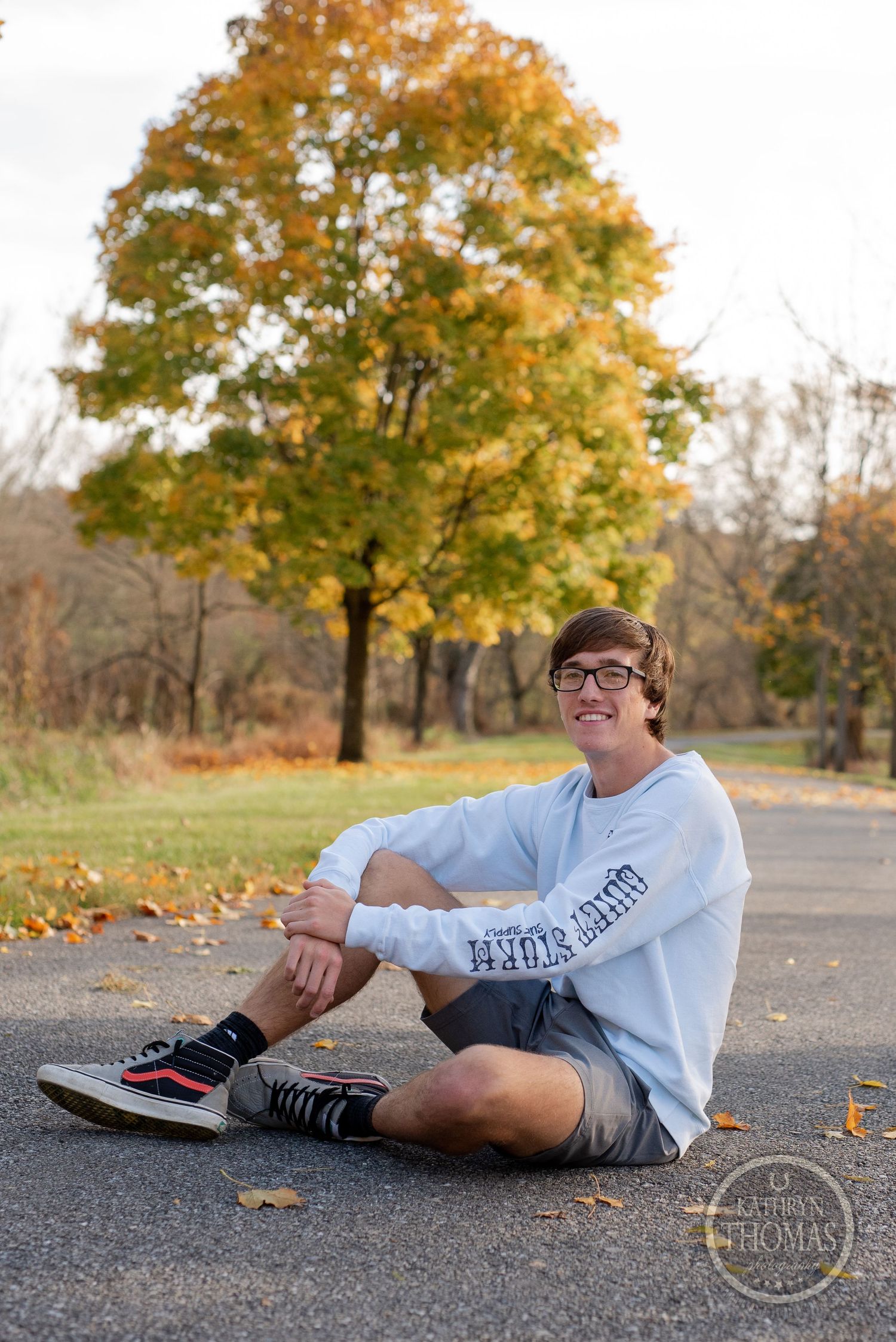 Person sits on autumn path wearing white sweatshirt and shorts with fall trees behind.