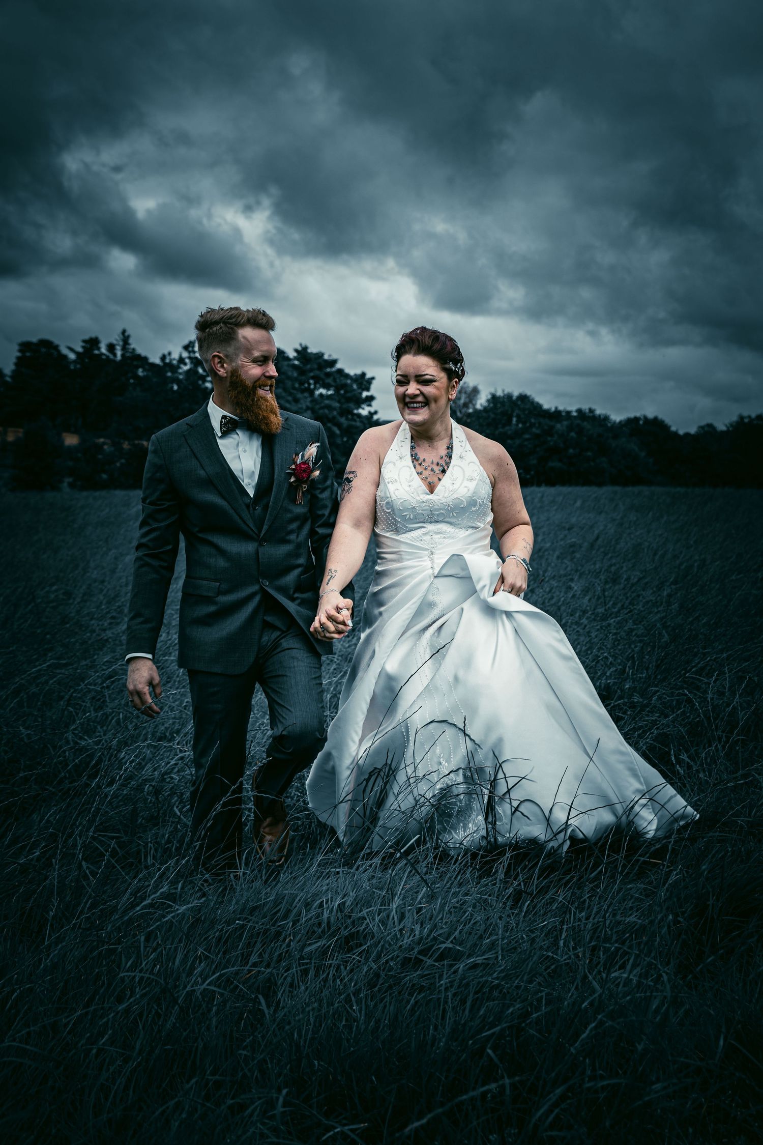 Laughing bride and bearded groom walking through dark field under stormy sky.