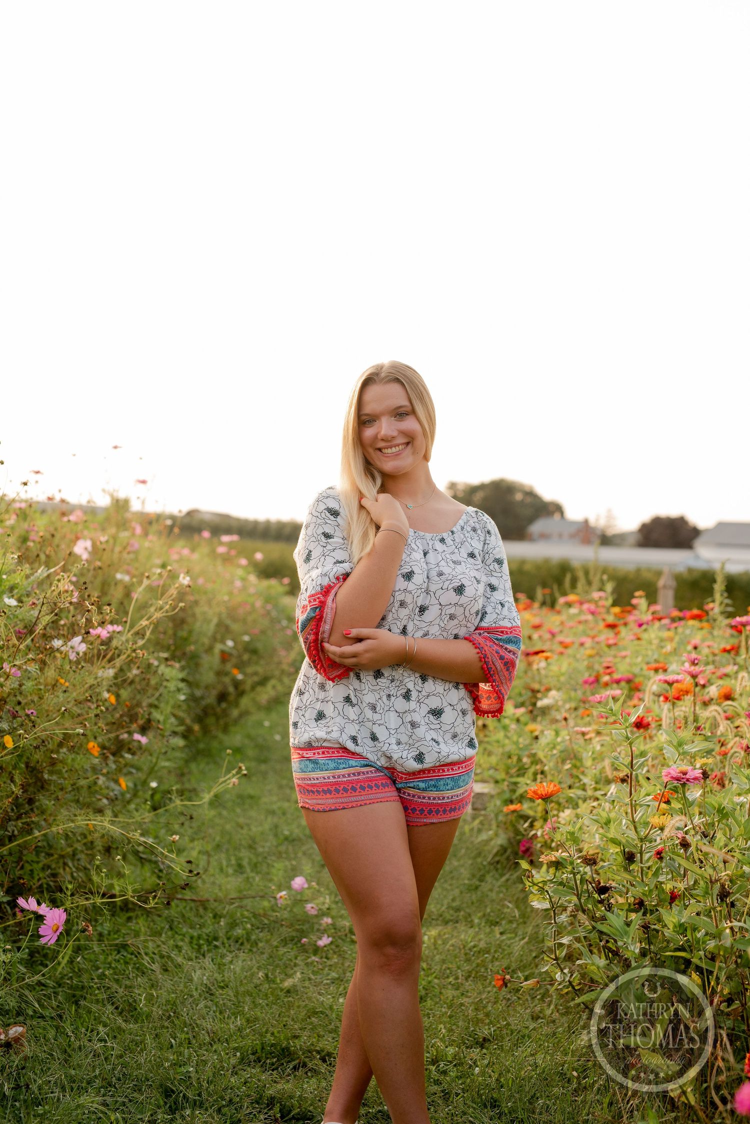 Standing in patterned summer outfit surrounded by wild poppies and flowers at sunset.