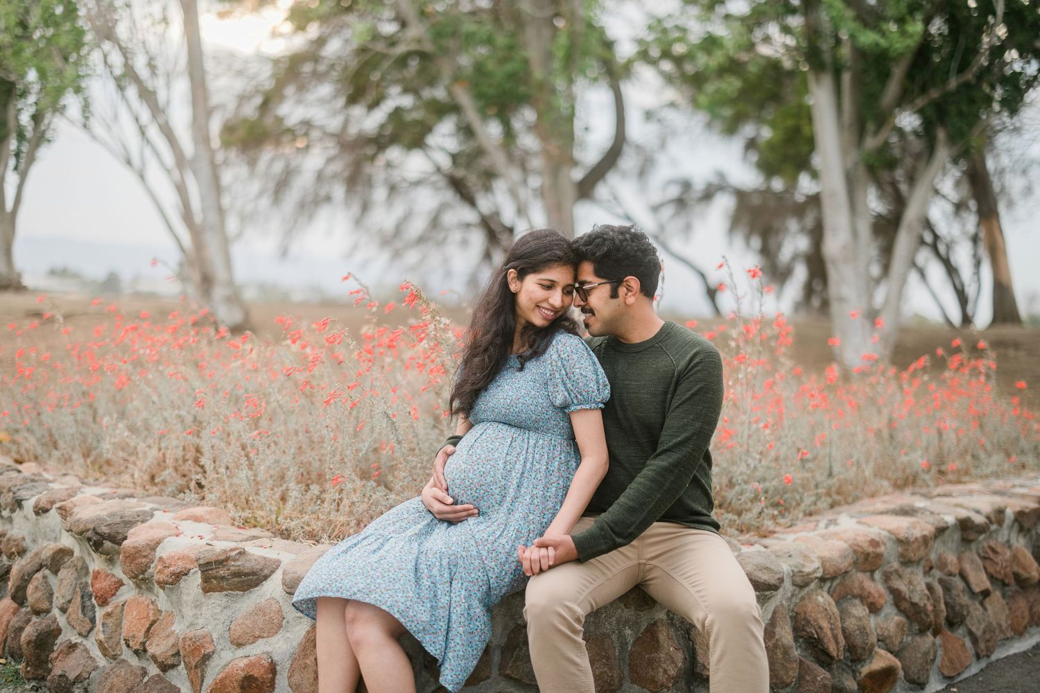 A couple sits together on a stone wall surrounded by red wildflowers and trees.