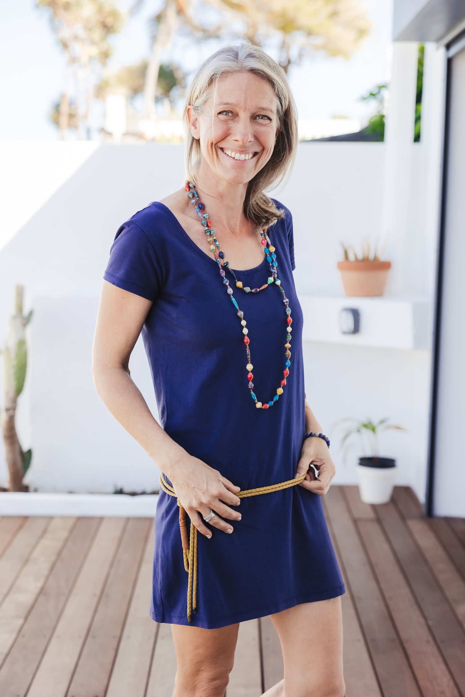 Woman in navy blue dress with colorful necklace posing playfully on wooden deck.