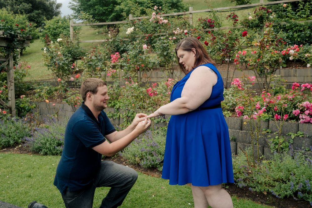 man in blue  shirt placing engagement ring on finger of plus size girlfriend in blue dress after surprise proposal