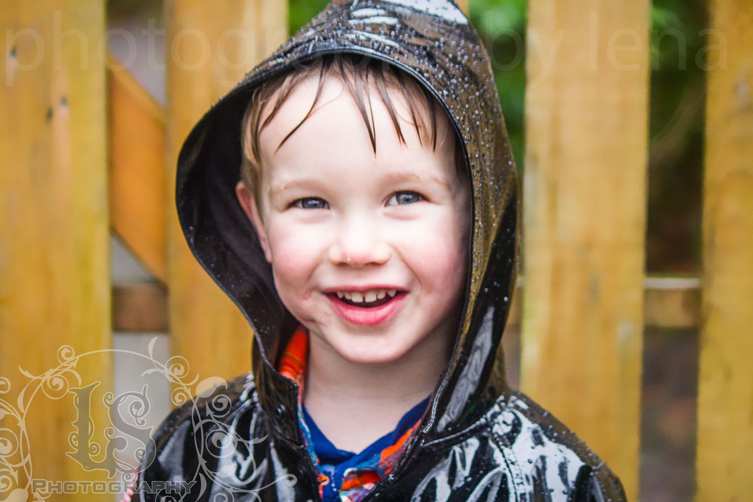 Small child wearing a black raincoat with hood smiles brightly in front of wooden fence.