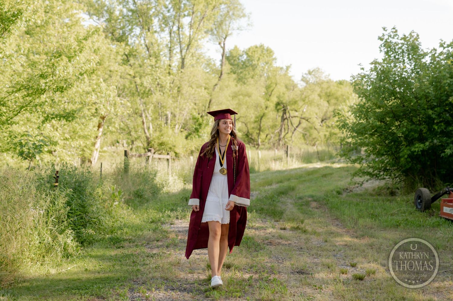 Graduate in maroon cap and gown walking on outdoor path surrounded by green trees.