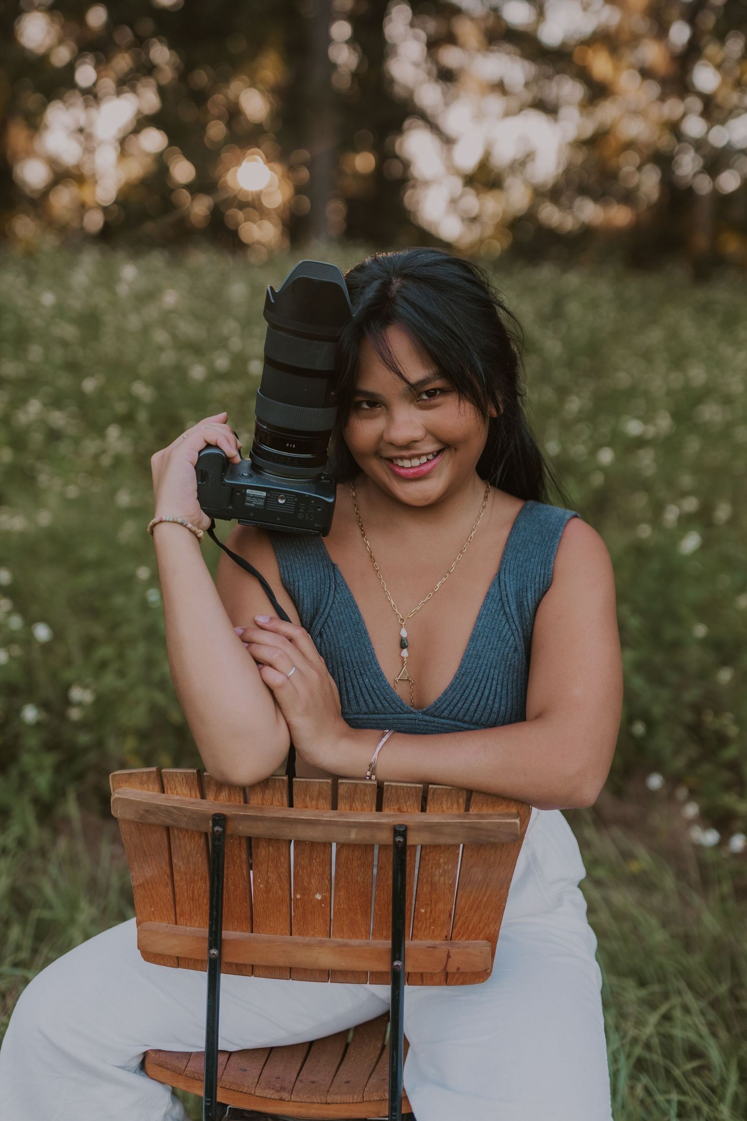 Photographer poses with professional camera while sitting on wooden chair in sunlit garden setting.