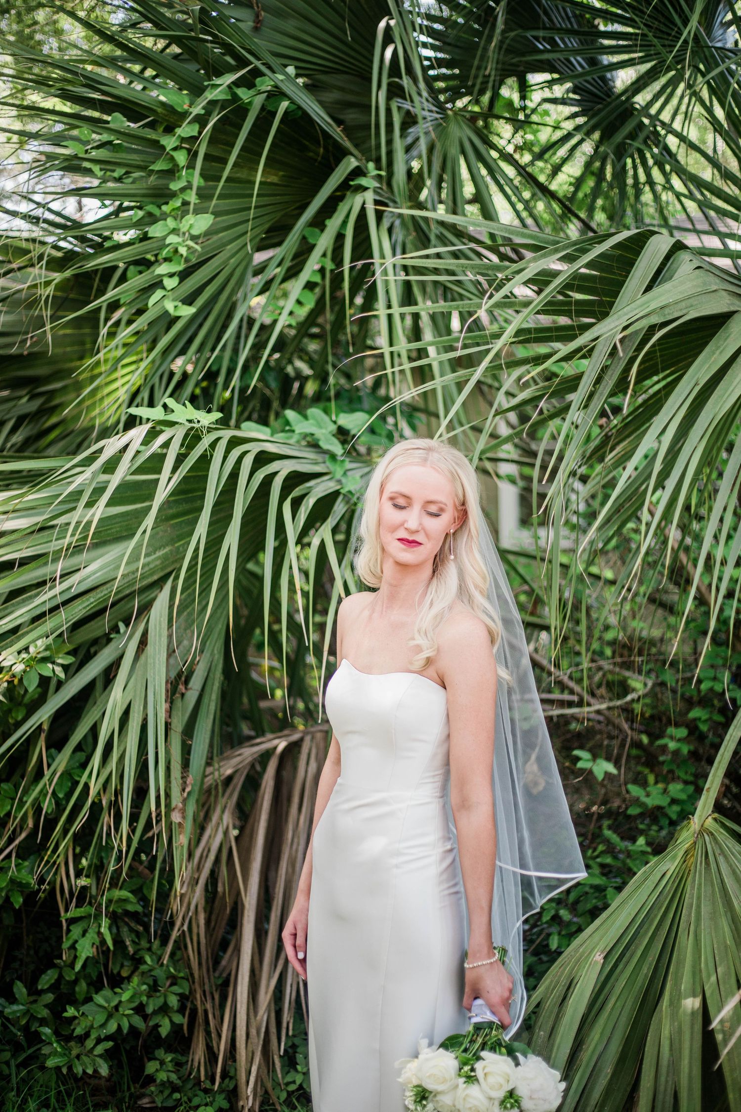 A bride in an elegant white gown poses among lush tropical palm leaves.