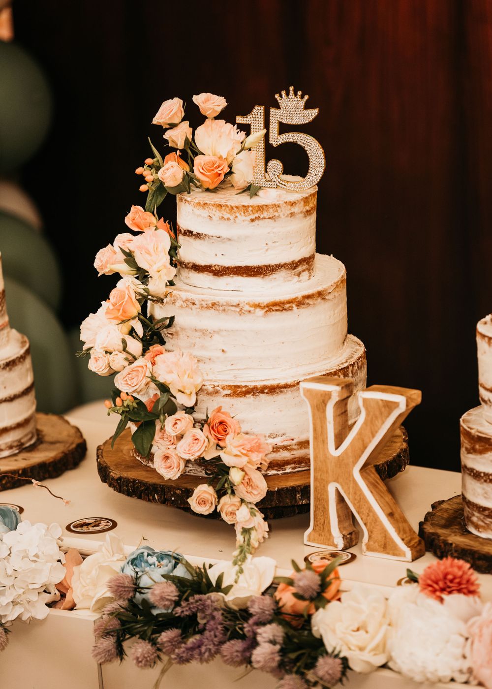 Three-tiered semi-naked wedding cake decorated with peach flowers and letter K topper.