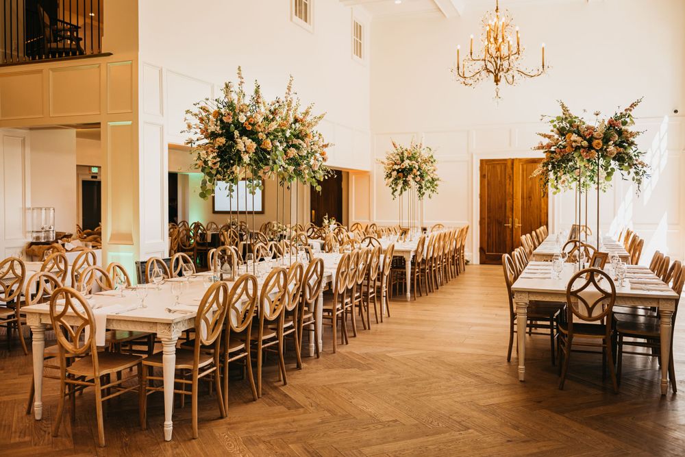 Elegant wedding reception venue with long dining tables, wooden chairs and tall floral centerpieces under a chandelier.