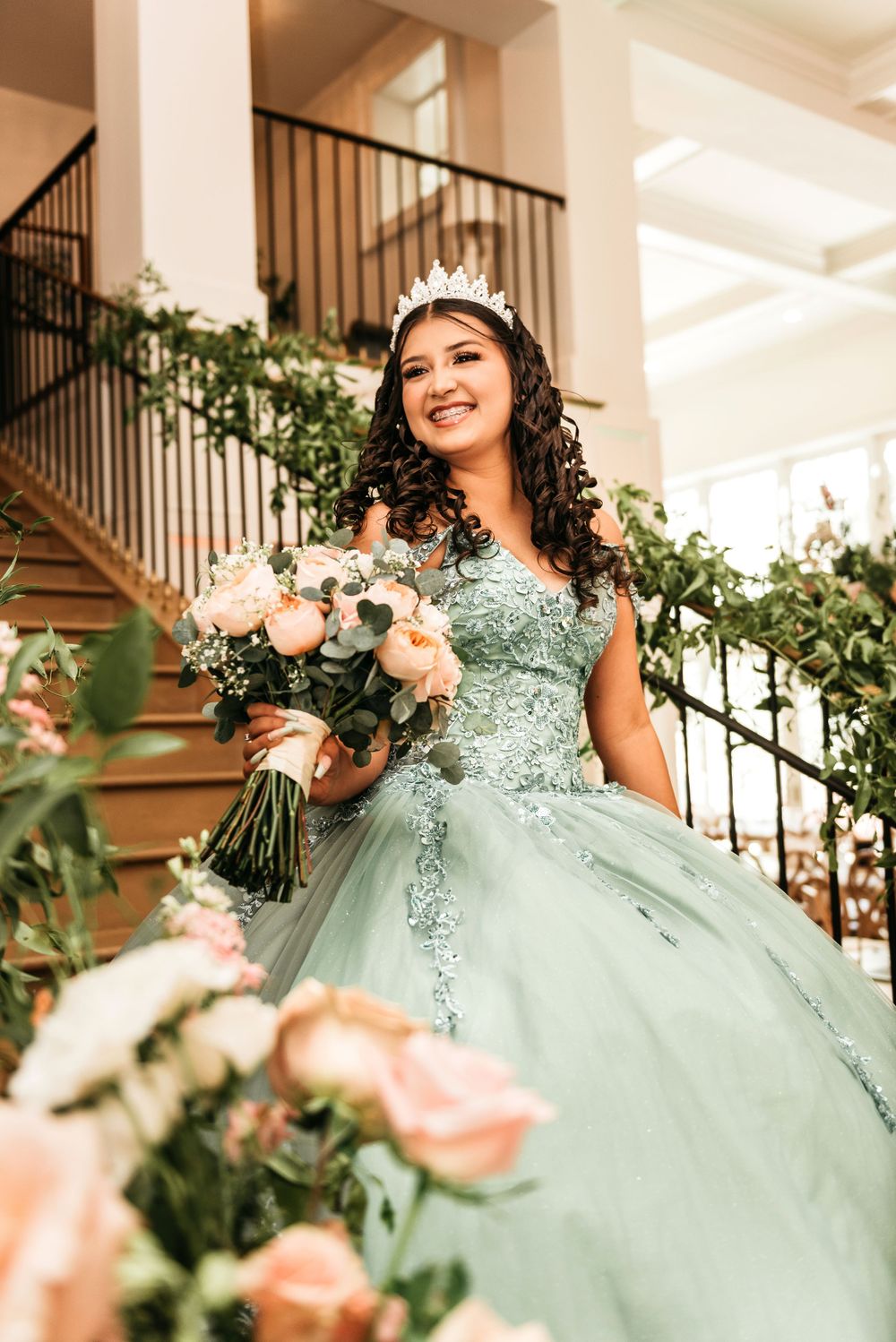 Person in mint green embellished ballgown holding bouquet on elegant staircase.