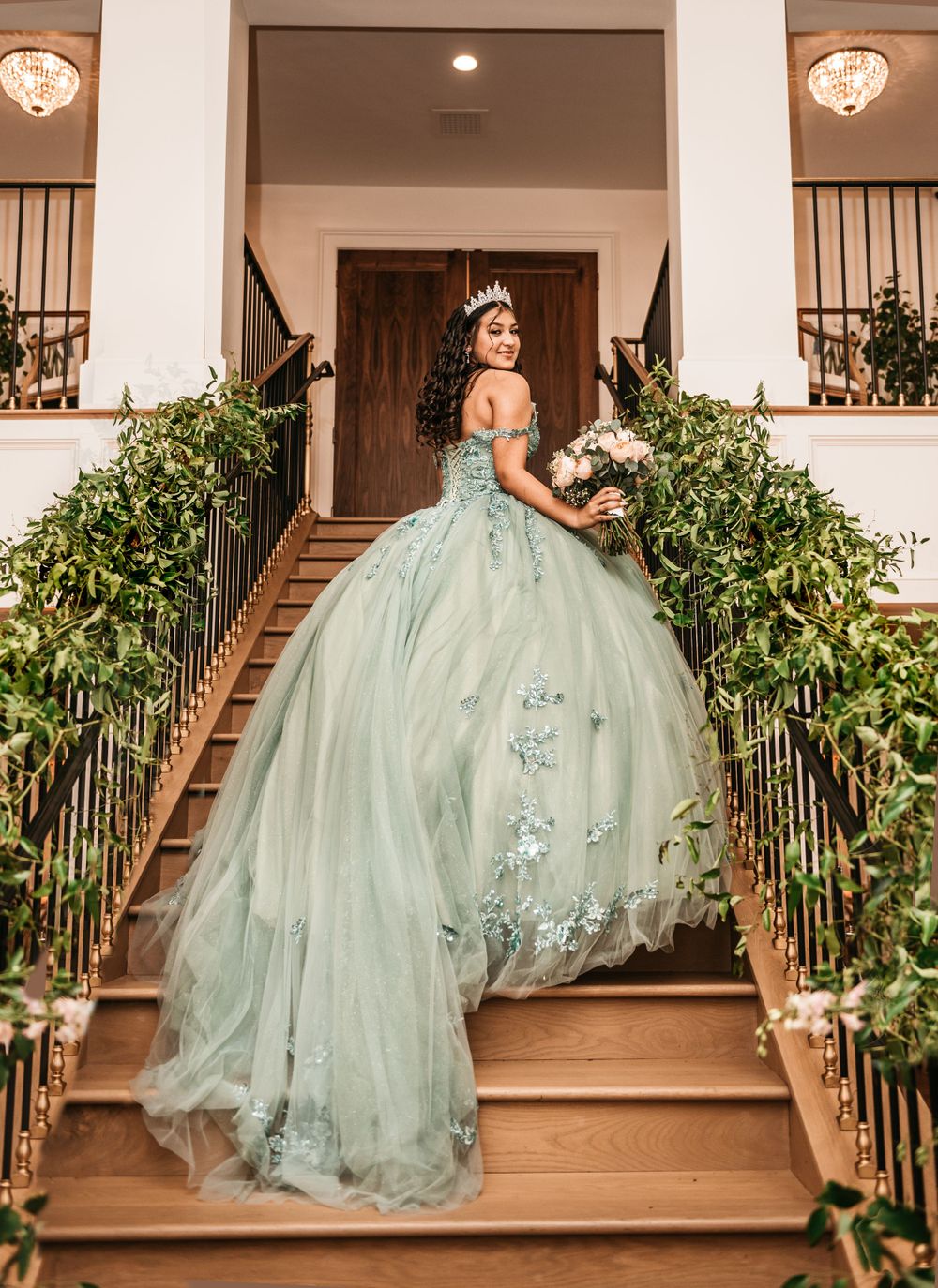 A mint green quinceañera ball gown with embellishments photographed on an elegant staircase adorned with greenery.