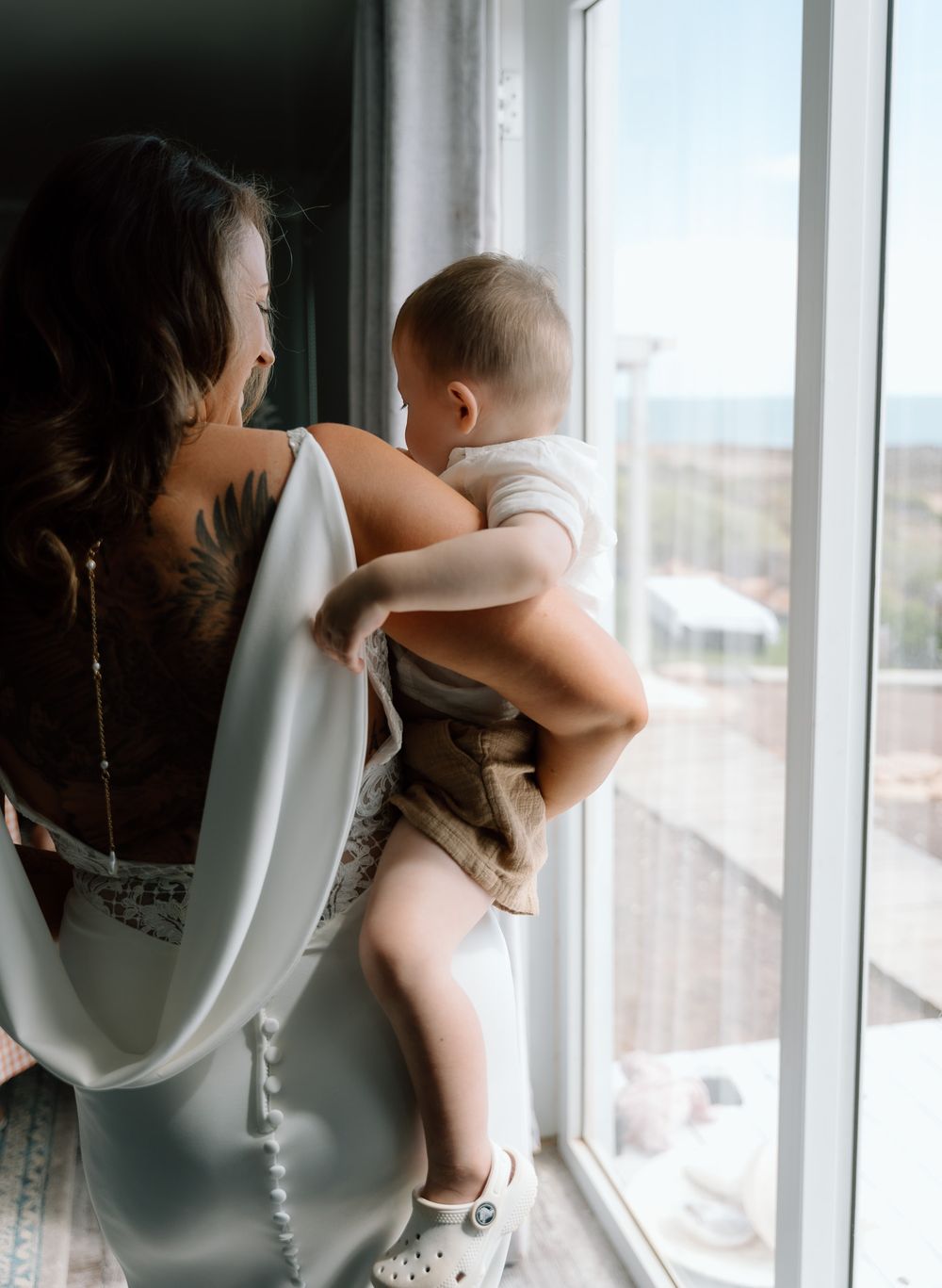 Parent and baby share an intimate moment by a sunlit window in elegant white attire.