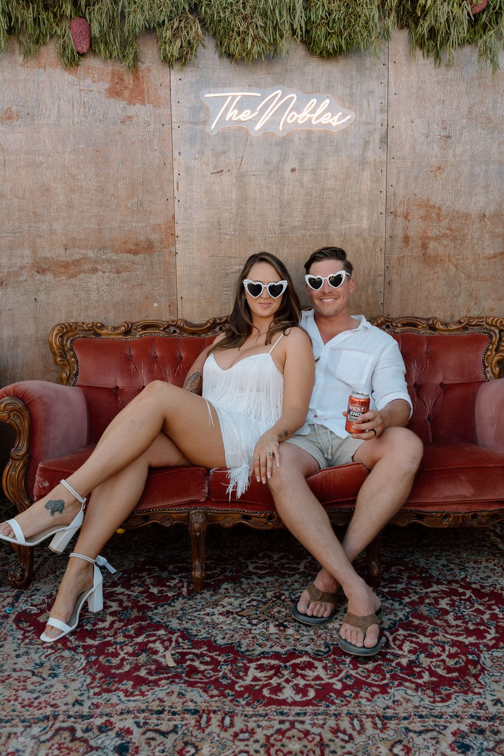 Couple relaxing on a vintage red couch with drinks at an outdoor venue.