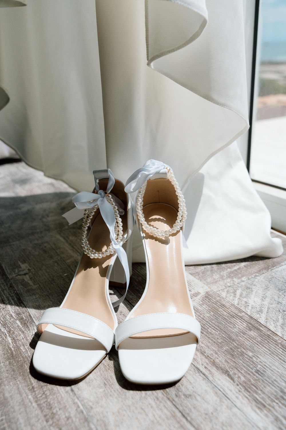Elegant white wedding shoes with pearl ankle straps displayed against a flowing dress.