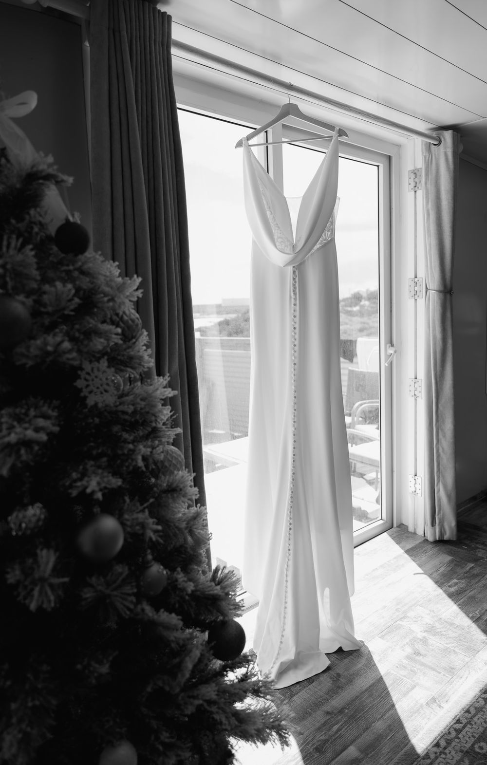 Elegant white wedding dress hanging by a window with a Christmas tree in the corner.