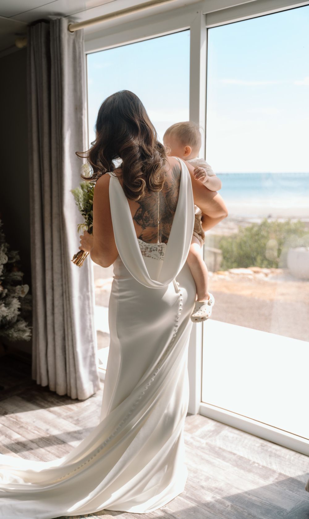 A tender moment between parent and child in white dress looking out a bright window with ocean views.