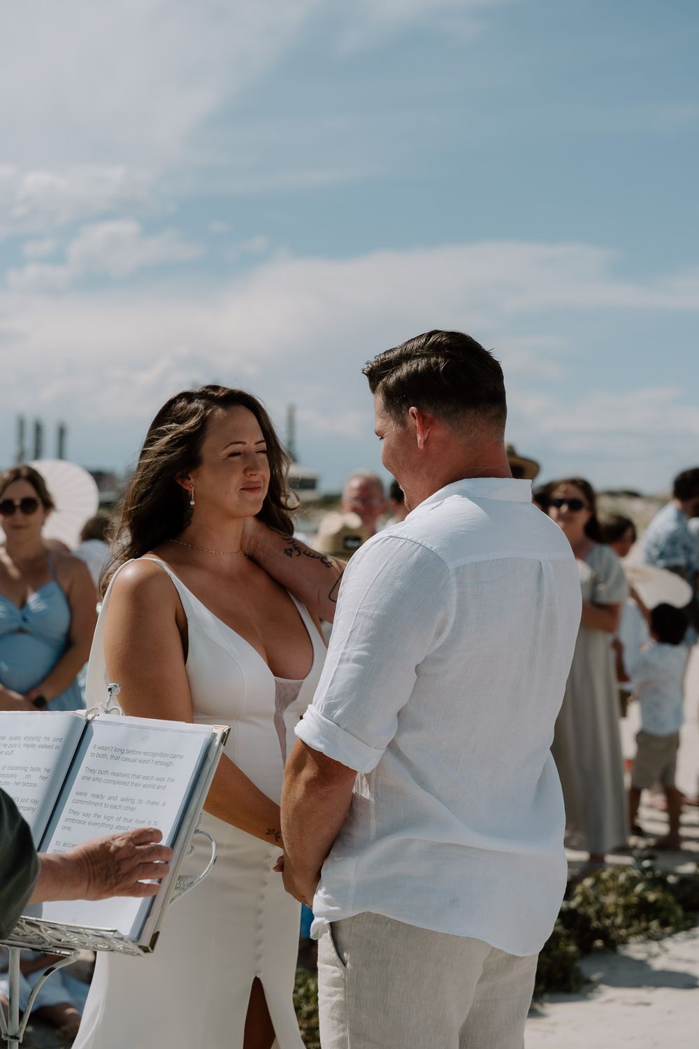 Wedding guests in white and beige attire gather on a sunny beach during an outdoor ceremony.