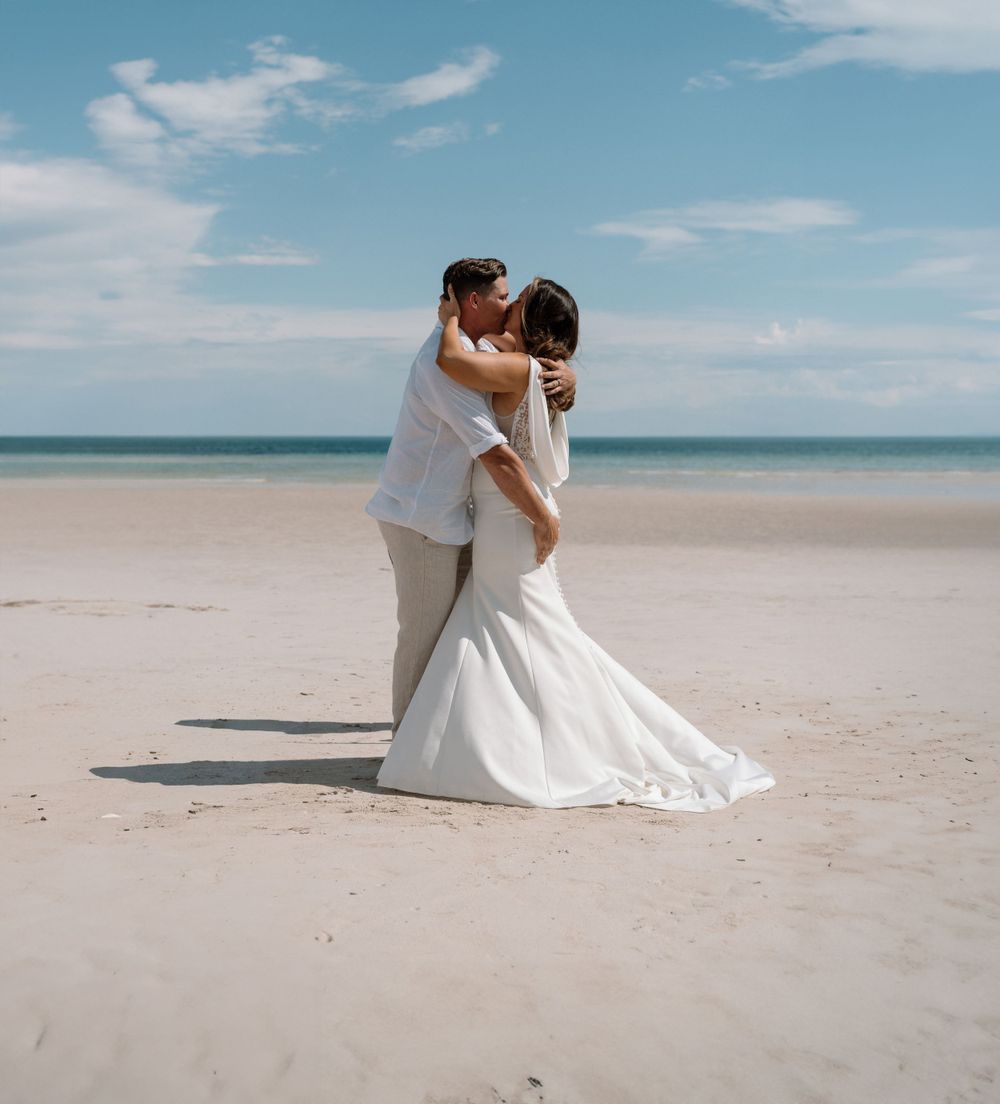 Newlyweds embracing on a sunny beach with white sand and blue skies.