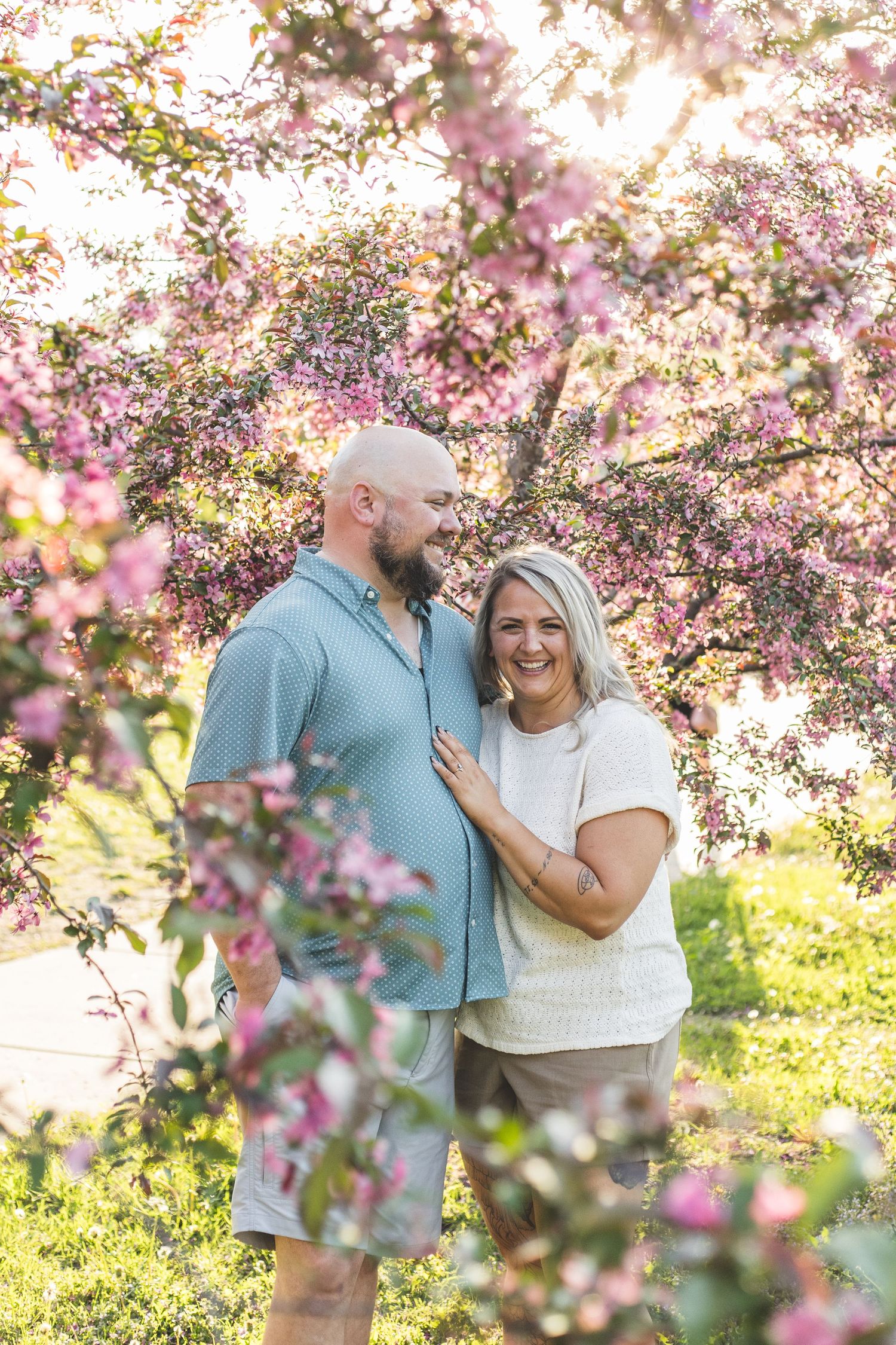 Couple smiling together under blooming pink cherry blossom trees in a sunny outdoor setting.
