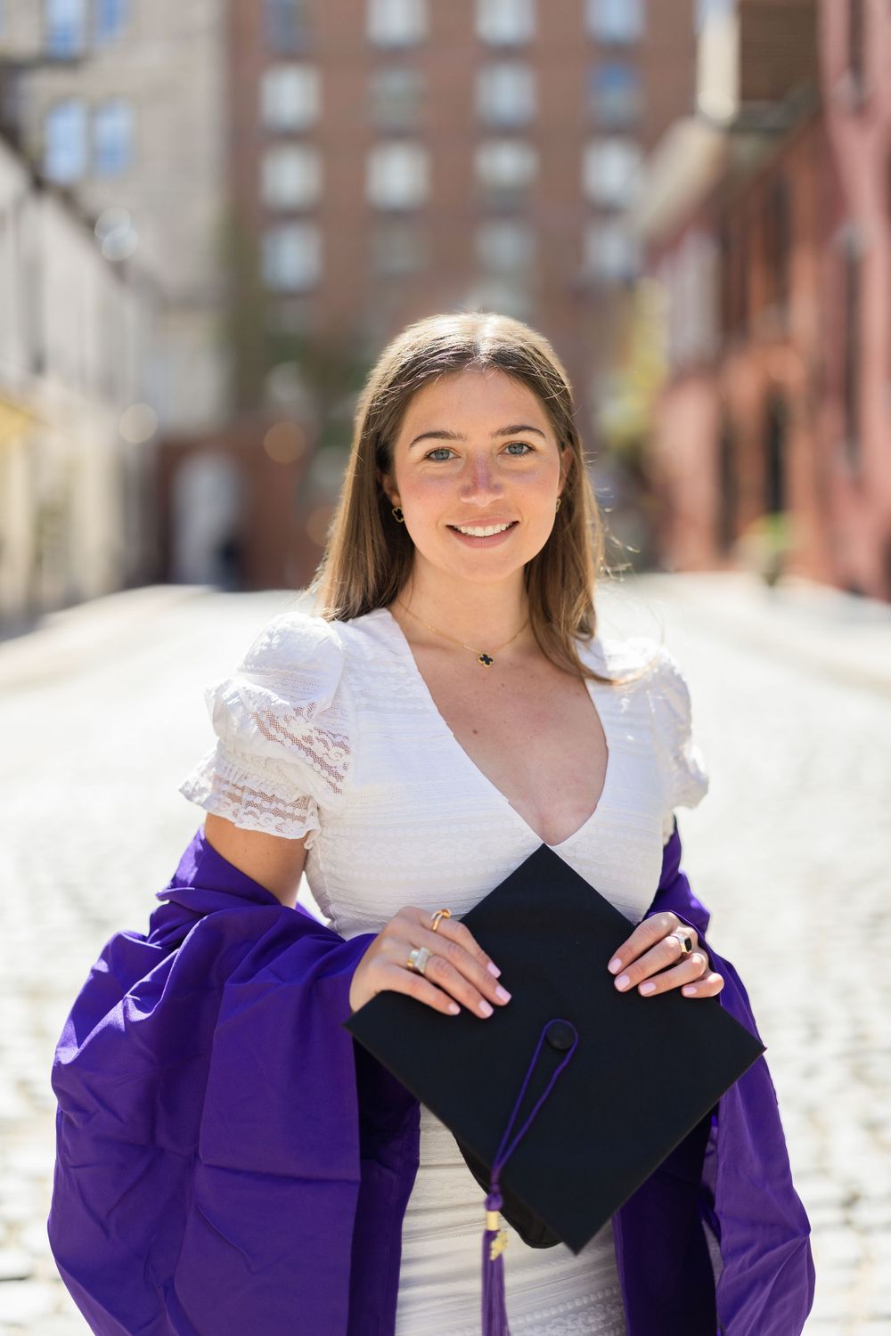 Graduate in white dress and purple regalia holds diploma on urban street.