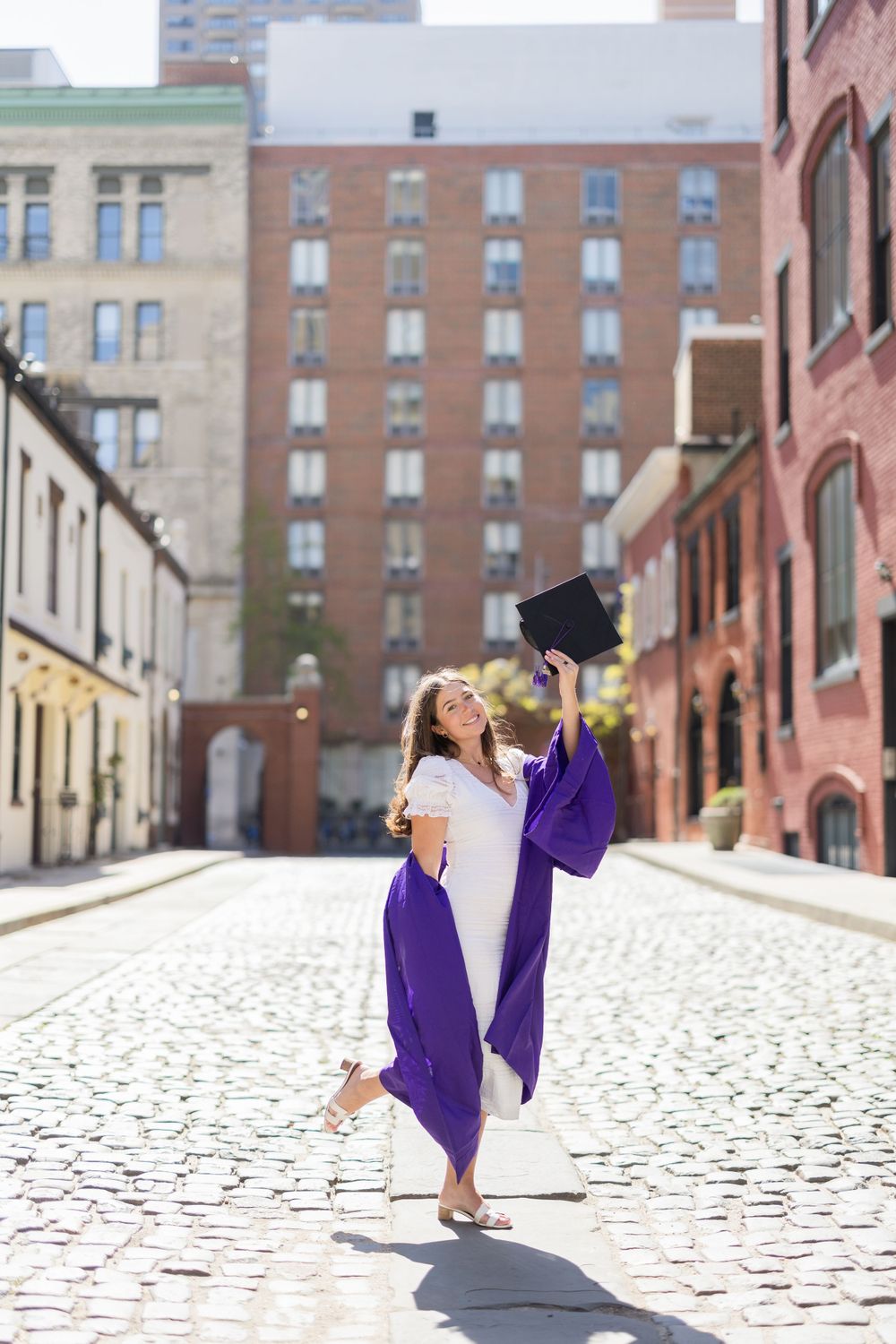 Graduate in purple gown jumps joyfully on cobblestone street.
