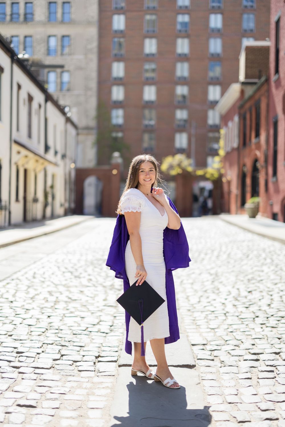 Graduate poses on cobblestone street holding cap and wearing purple gown.