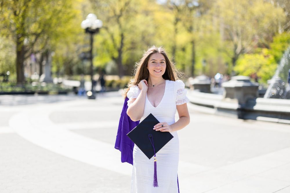 Graduate in white dress and purple gown poses in sunny park setting.