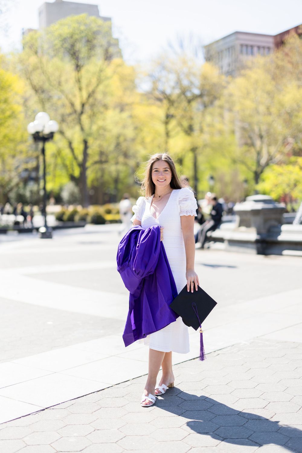 Graduate stands in spring park wearing white dress and holding purple graduation gown.