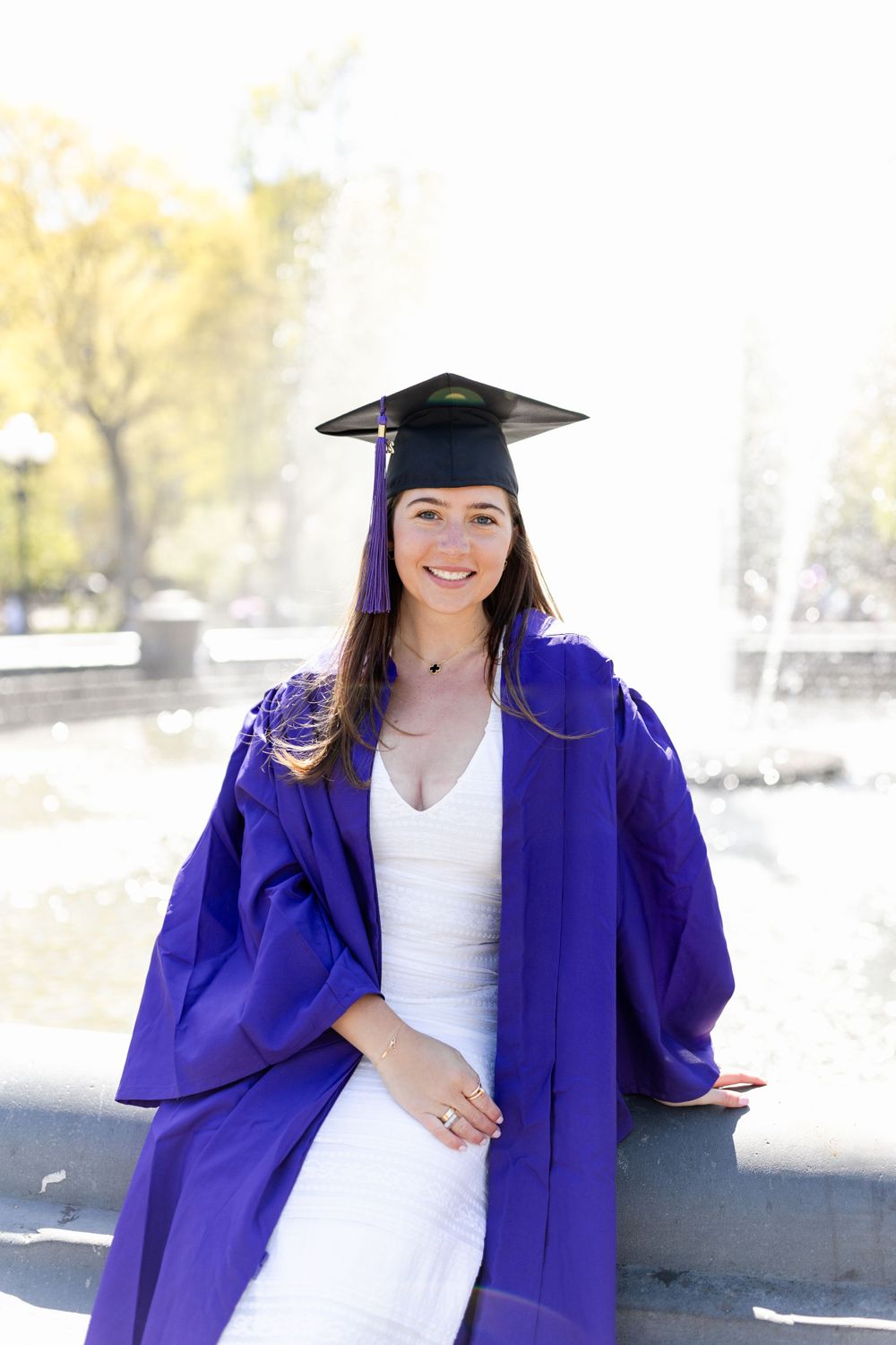 Graduate in purple academic regalia and cap poses outdoors with a fountain in background.