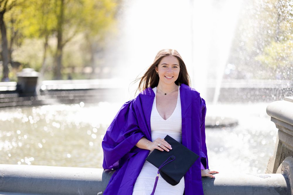 Graduate in white dress and purple graduation gown holds diploma case by fountain.