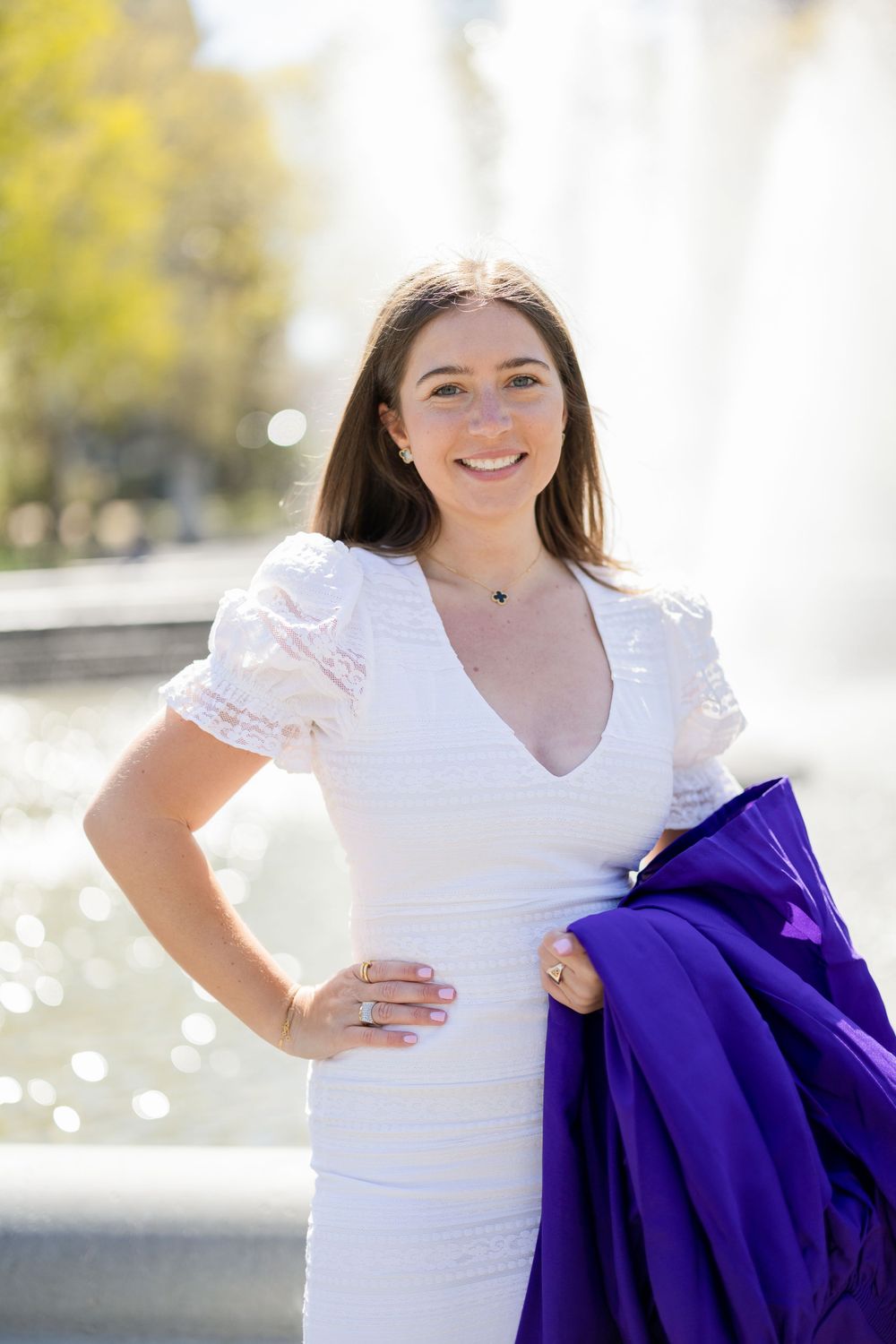 Graduate in white dress with purple academic gown poses near fountain.