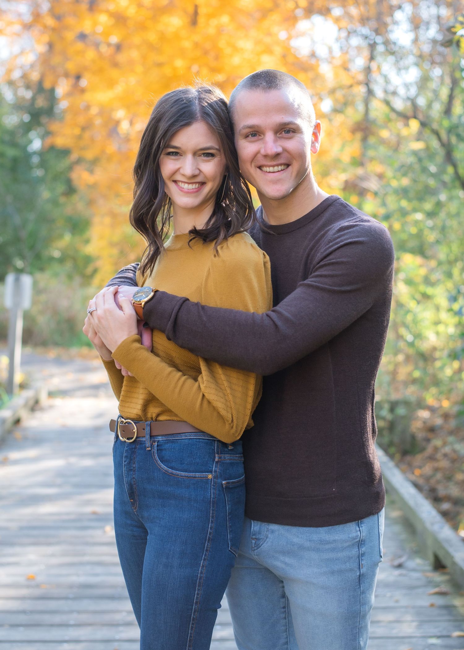 A couple embraces on a wooden walkway surrounded by fall foliage.