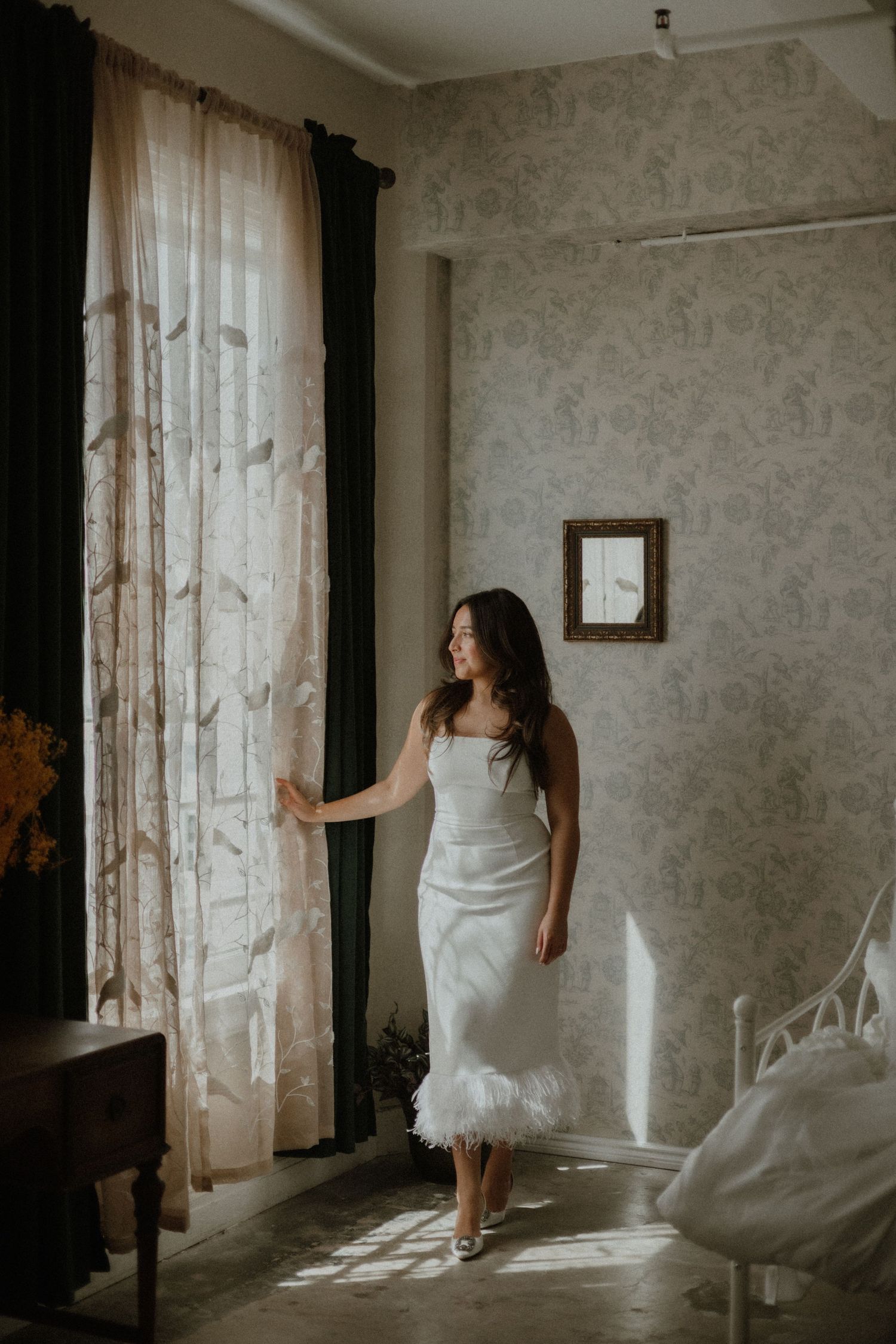 A figure in a white dress standing by a window with sheer curtains in a room with floral wallpaper.