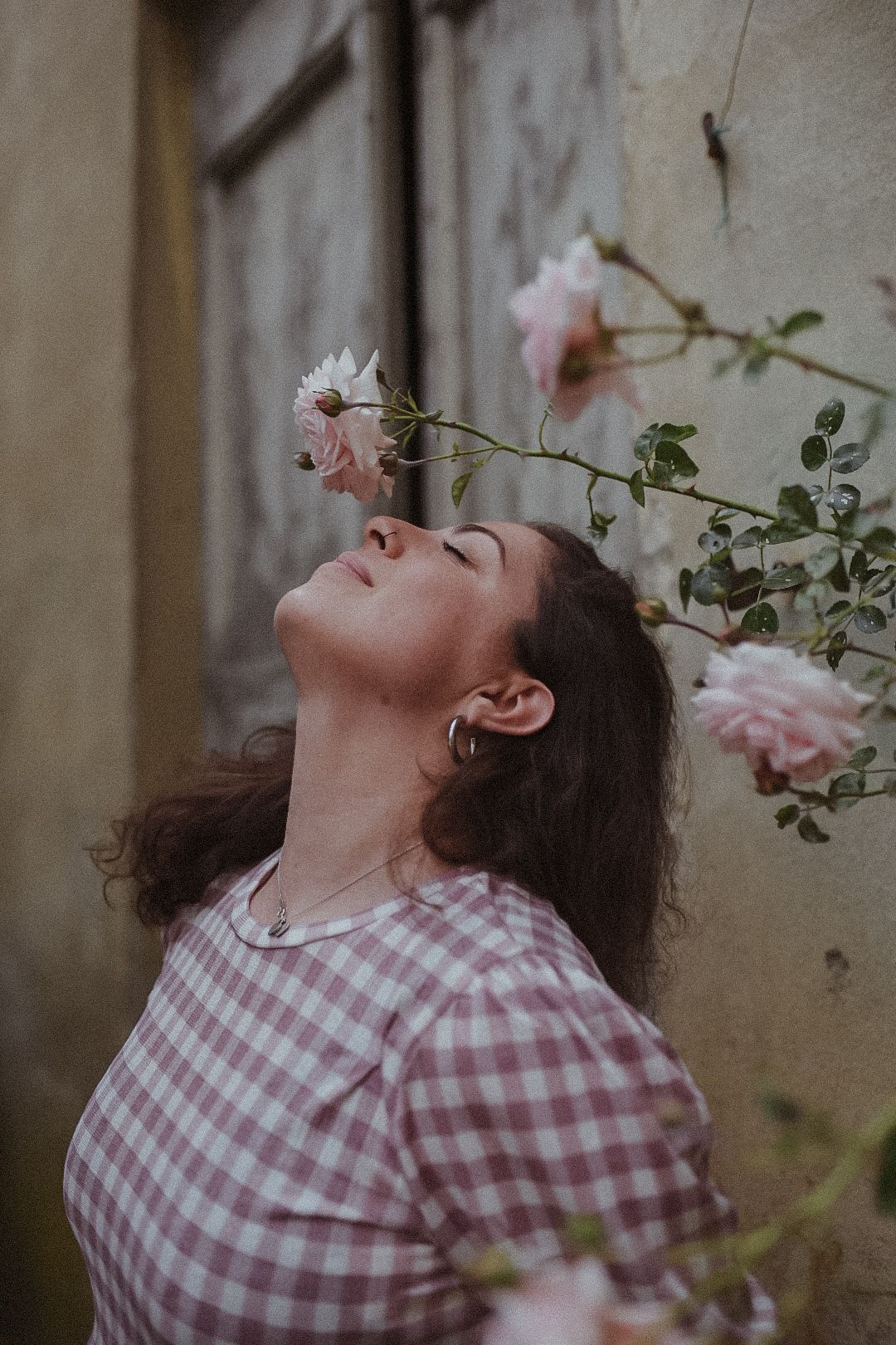 Person in pink gingham shirt poses artistically with delicate pink roses in a garden setting.