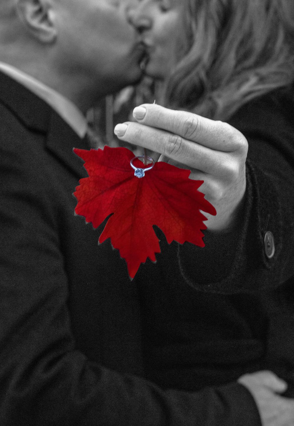 Bride and groom kissing in a blurred background holding a red leaf with engagement ring