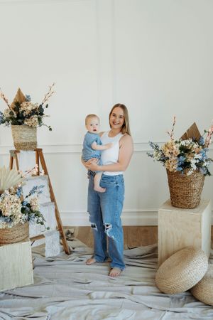 Mother and baby pose with rustic floral decorations and baskets in a bright studio setting.