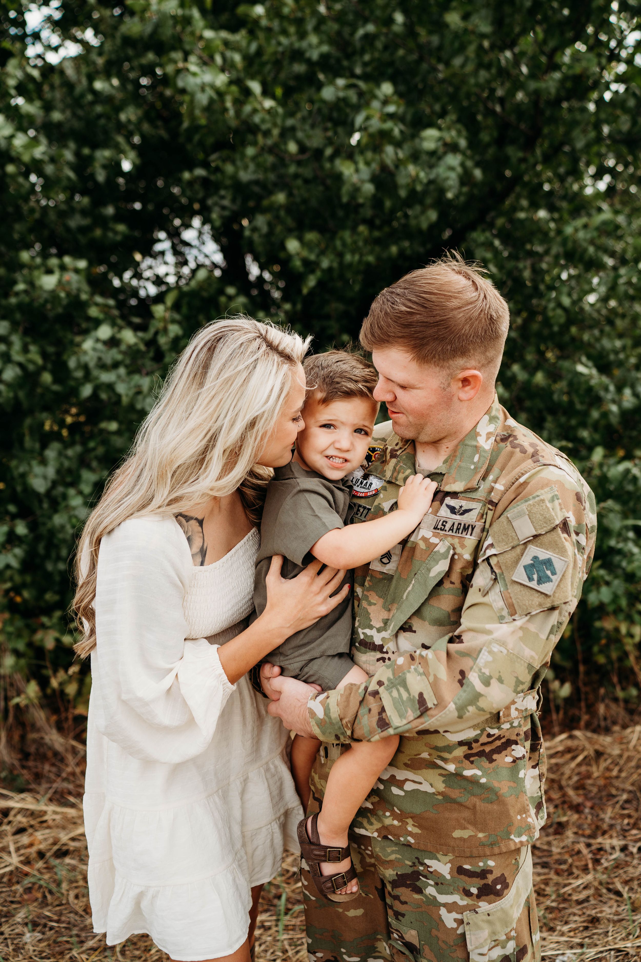 Military family embraces outdoors with toddler in father's arms showing tender moment.