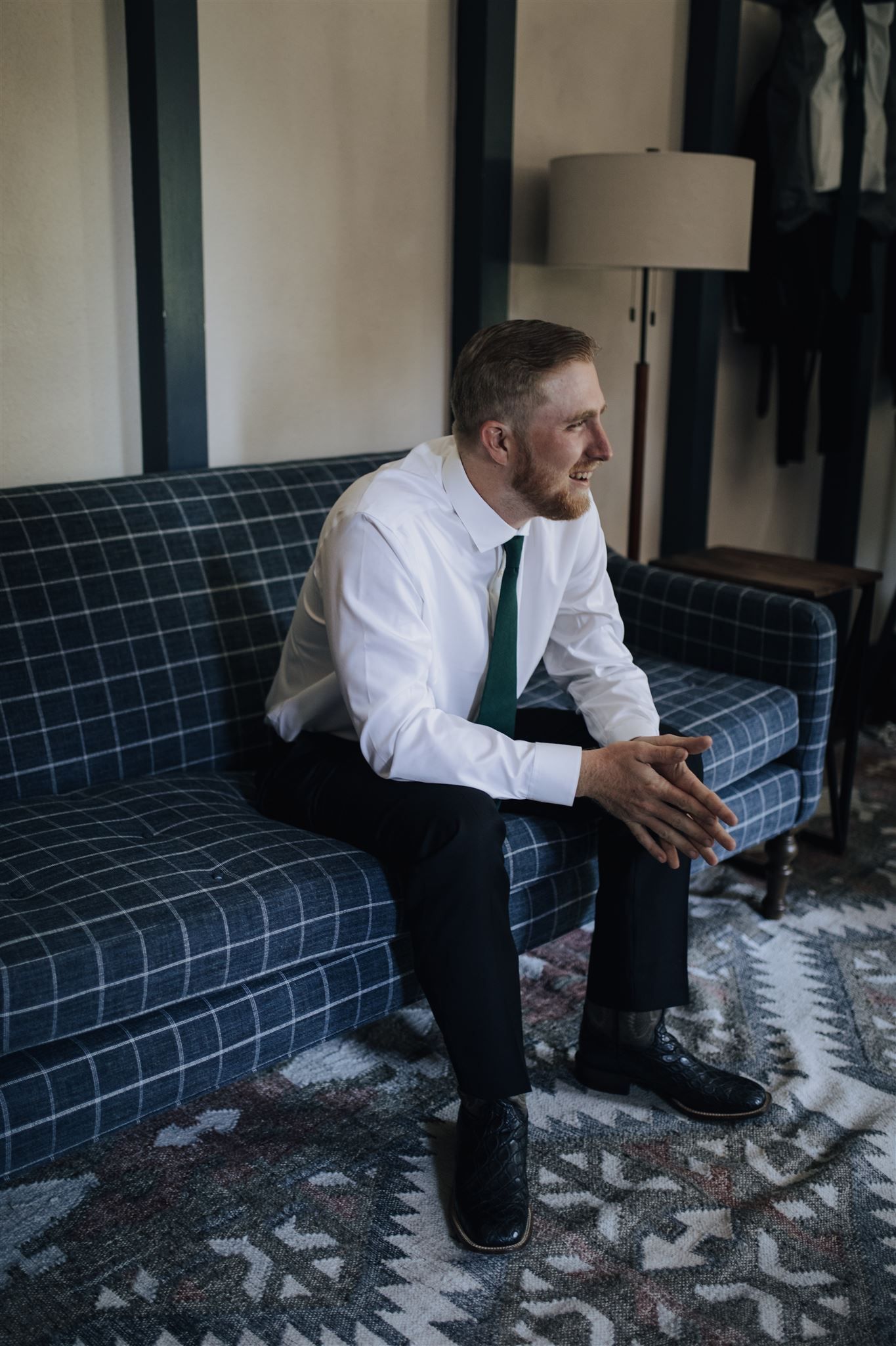 Man in formal attire sits pensively on a blue plaid couch before wedding ceremony.