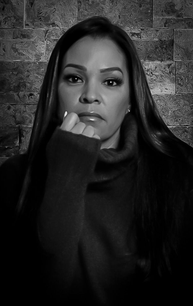 Black and white portrait of a woman with long hair resting her chin on her hand against a brick wall.