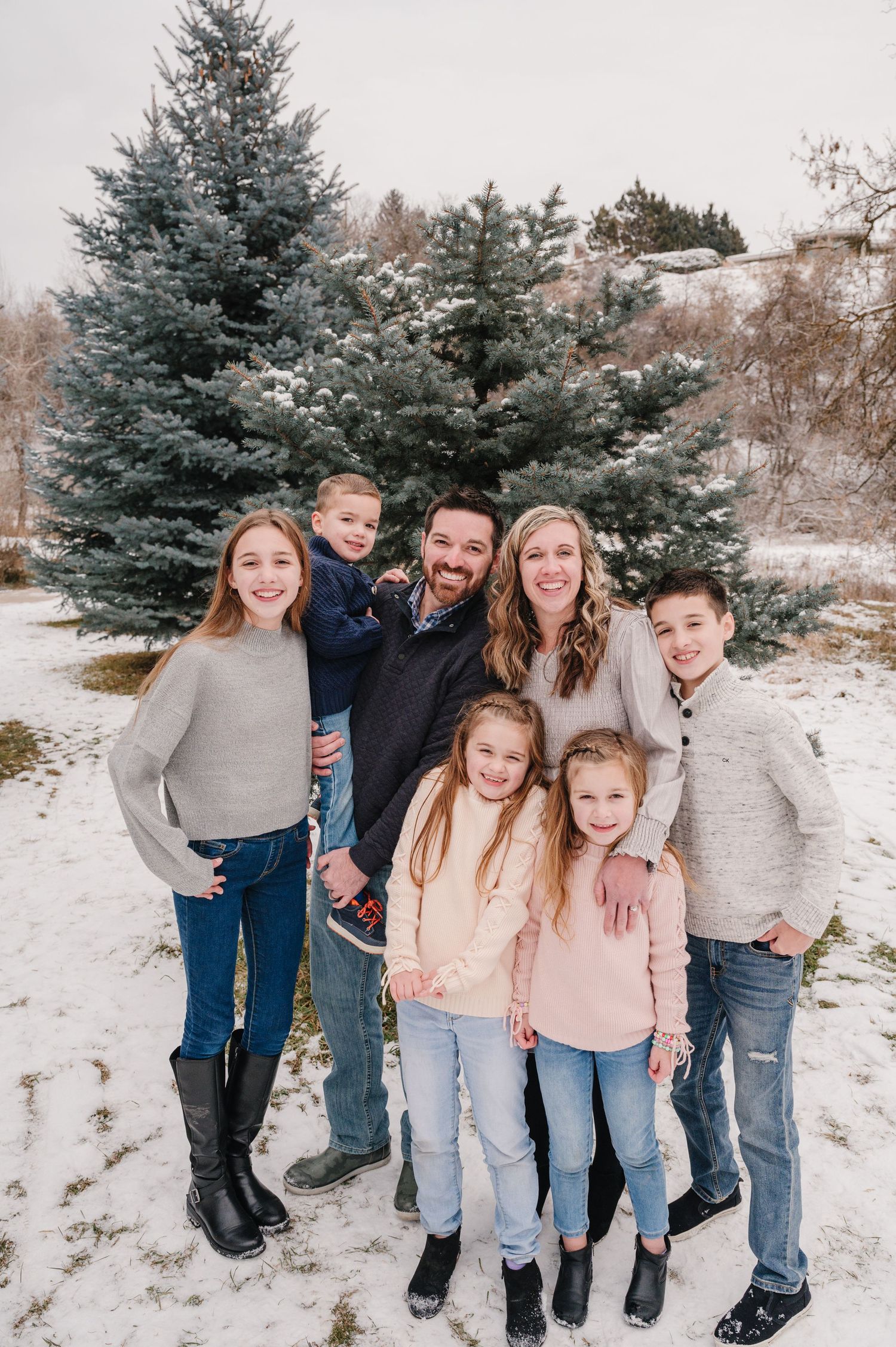 Family group poses together in winter scene with snow-covered blue spruce trees.