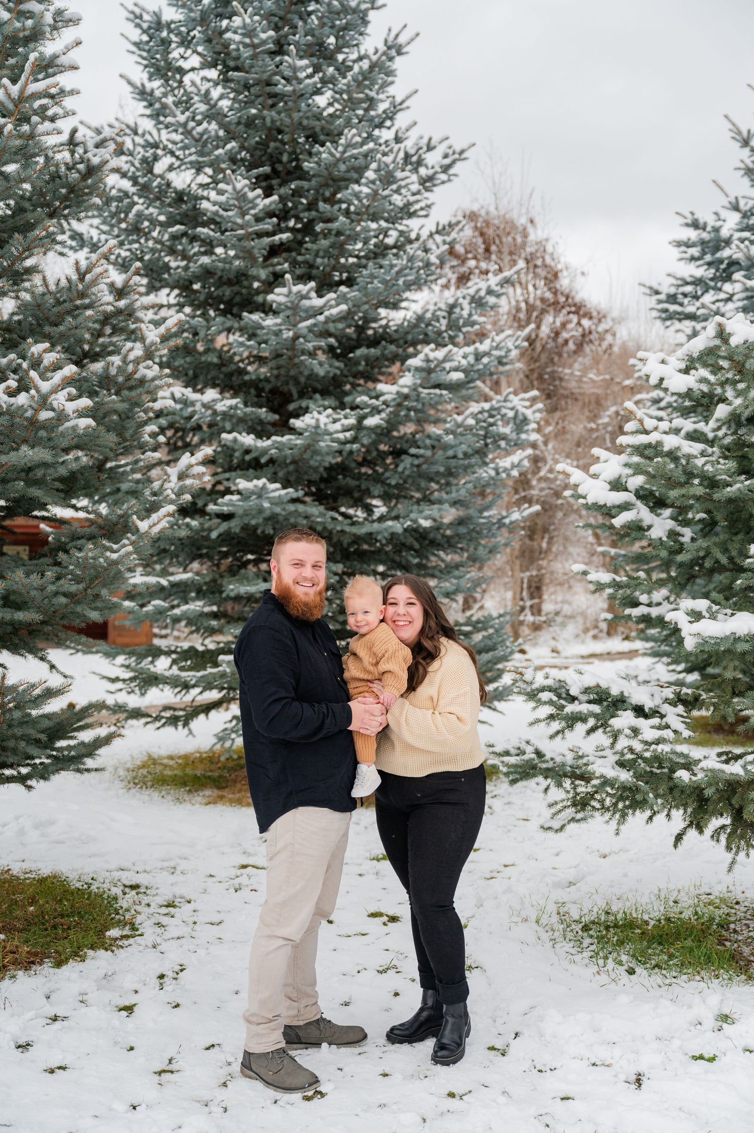 Small family posing together in front of snow-dusted evergreen trees during winter.