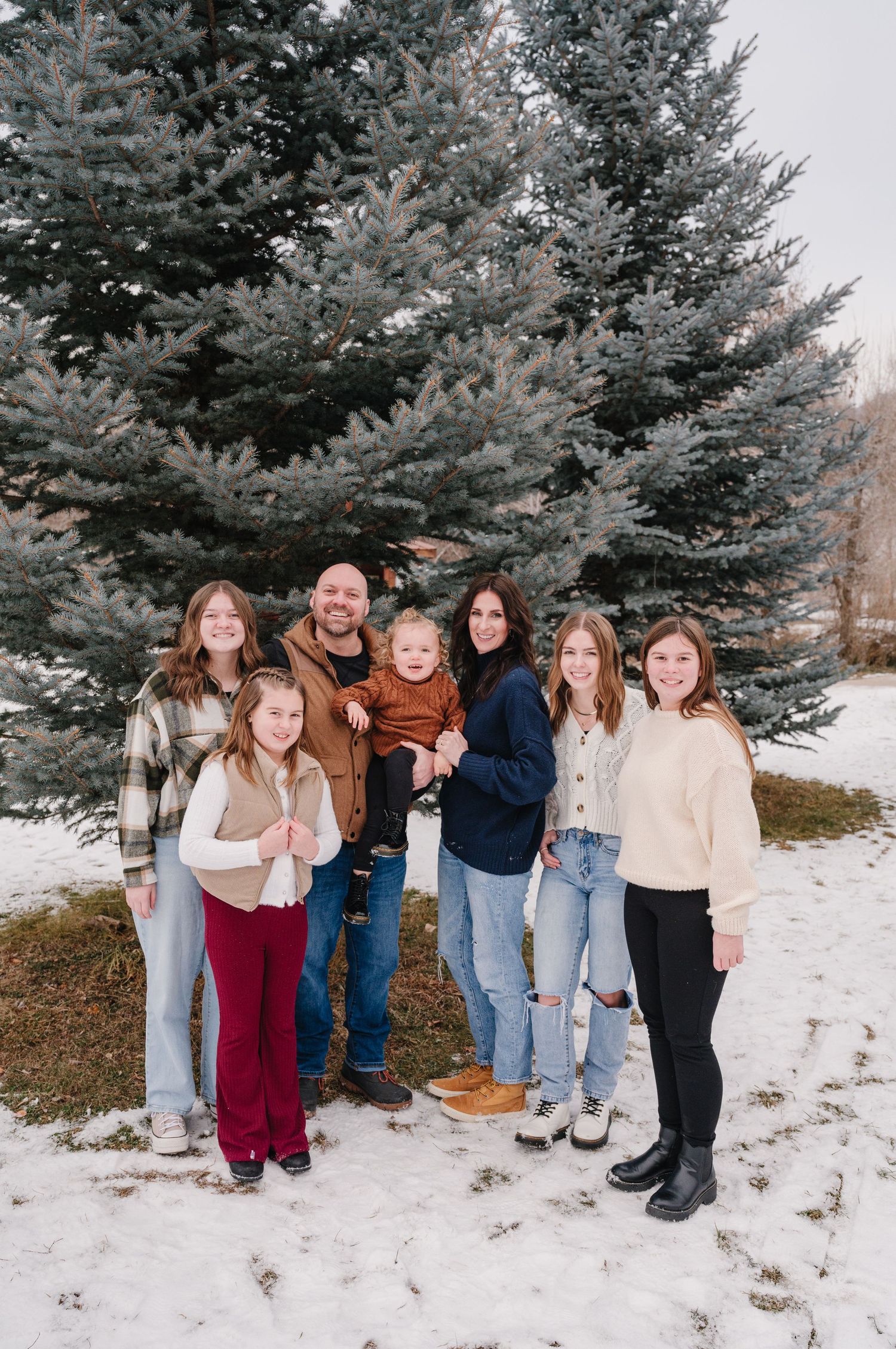 A group stands together in winter against tall blue spruce trees in the snow wearing casual winter attire.