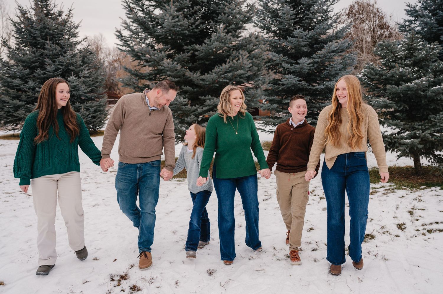 Five people walking hand in hand through snowy landscape with evergreen trees in background.